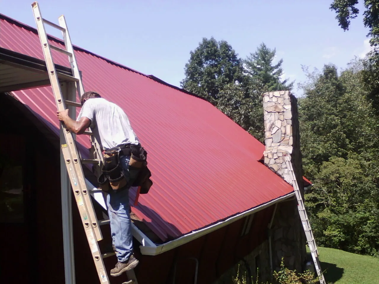 Roofer on a ladder beside a red metal roof and stone chimney — Soffit & Fascia Repair by Roofing Brothers in Votaw