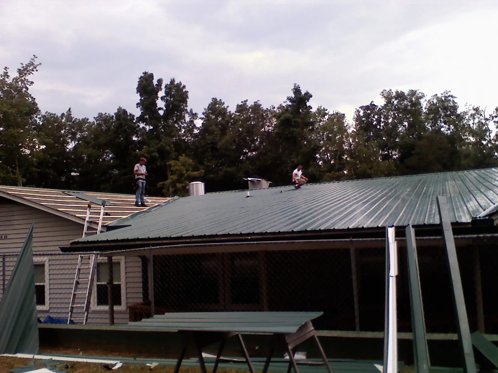 Two roofers installing green metal panels above a covered porch in Votaw